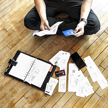 IMAGE: Person sitting on floor holding credit card and receipts with more receipts, cards, and a planner scattered on floor