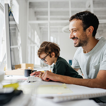 IMAGE: Man sitting at computer with small boy on his lap watching his dad's hands on keyboard.