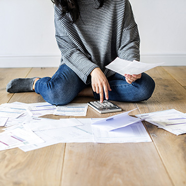 IMAGE: Below-shoulder view of woman sitting on floor with calculator among scattered papers