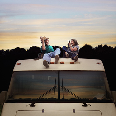 IMAGE: Mature couple reclined on roof of RV at sunset. Man posing for picture woman is taking with camera.