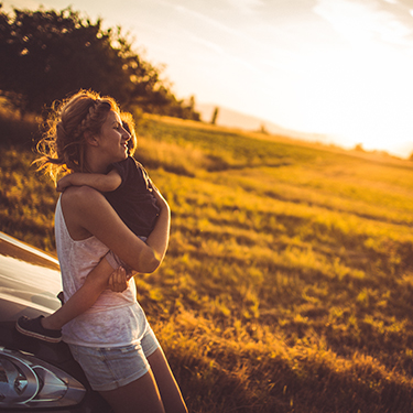 IMAGE: Woman hugging small child sitting on car hood with sunset behind