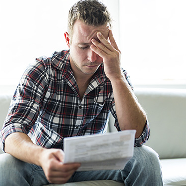 IMAGE: Man sitting with hand to forehead, looking concerned at some papers.