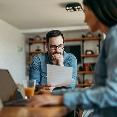 IMAGE: Man at table looking concerned at paper, woman in foreground looking towards him.