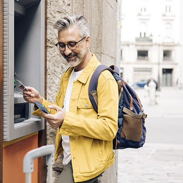 IMAGE: Man wearing backpack holding phone at ATM 