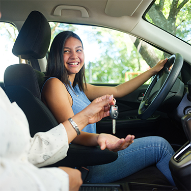 IMAGE: Female teen driver smiling behind wheel and being handed keys to the vehicle.