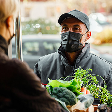 IMAGE: Man in protective mask delivering groceries to woman in doorway.