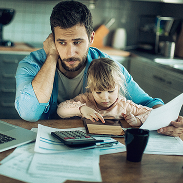 IMAGE: Worried man with young daughter in his lap at a table with wallet, papers, and a calculator