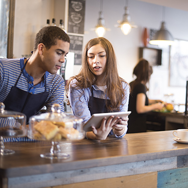 IMAGE: Two young adults working behind counter at coffee shop looking at tablet together