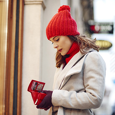 IMAGE: Woman outside with bright red winter hat, looking into her open wallet.