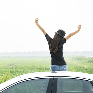 IMAGE: Girl sitting on car, facing away, with arms outstretched above in a celebratory pose.