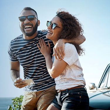 IMAGE: Man and woman smiling sitting on a car with ocean in the background