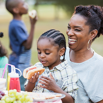 IMAGE: Mom laughing while child sits on her lap eating a hot dog at a backyard BBQ